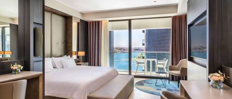 A modern hotel room with a large bed, a desk, a chair, a small table, and a balcony with a view of the sea.