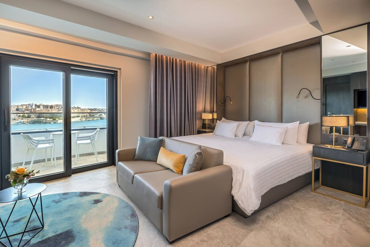A modern hotel room with a large bed, a sofa, a small table with a vase, and a balcony with a city view.