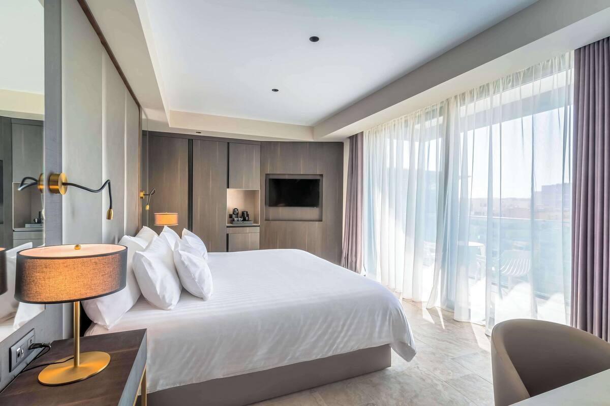A modern hotel room with a large bed, a desk with a lamp, a TV, and a balcony with curtains.