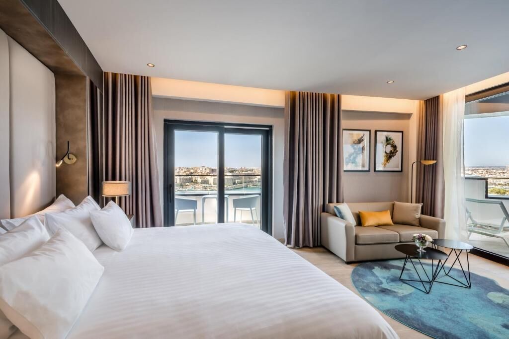 A hotel room with a large bed, a sofa, a small table, and a balcony with a city view.