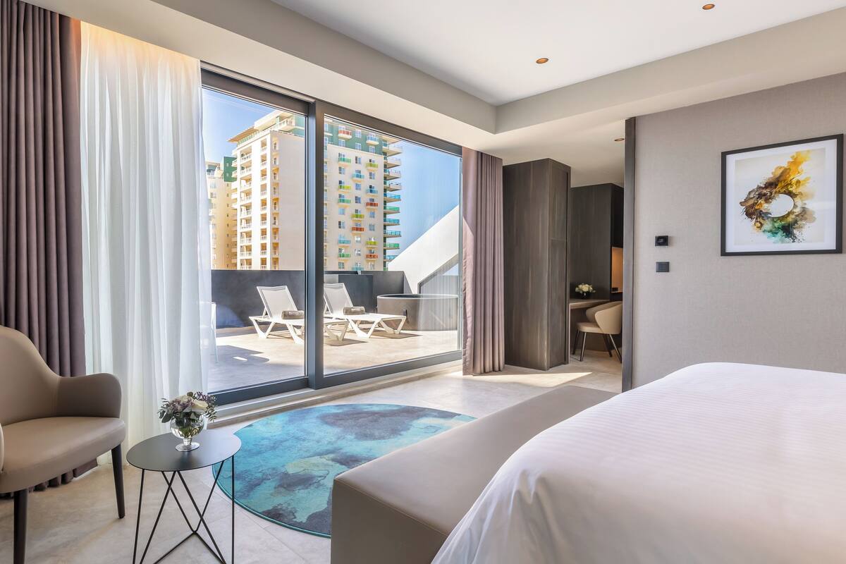A modern hotel room with a large bed, a seating area with chairs, a small table with a vase, and a view of a building through large windows.