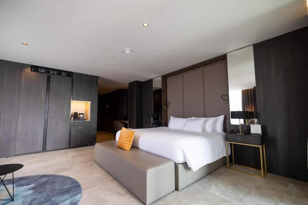 A modern hotel room with a large bed, a seating area, a desk, and a mirror.