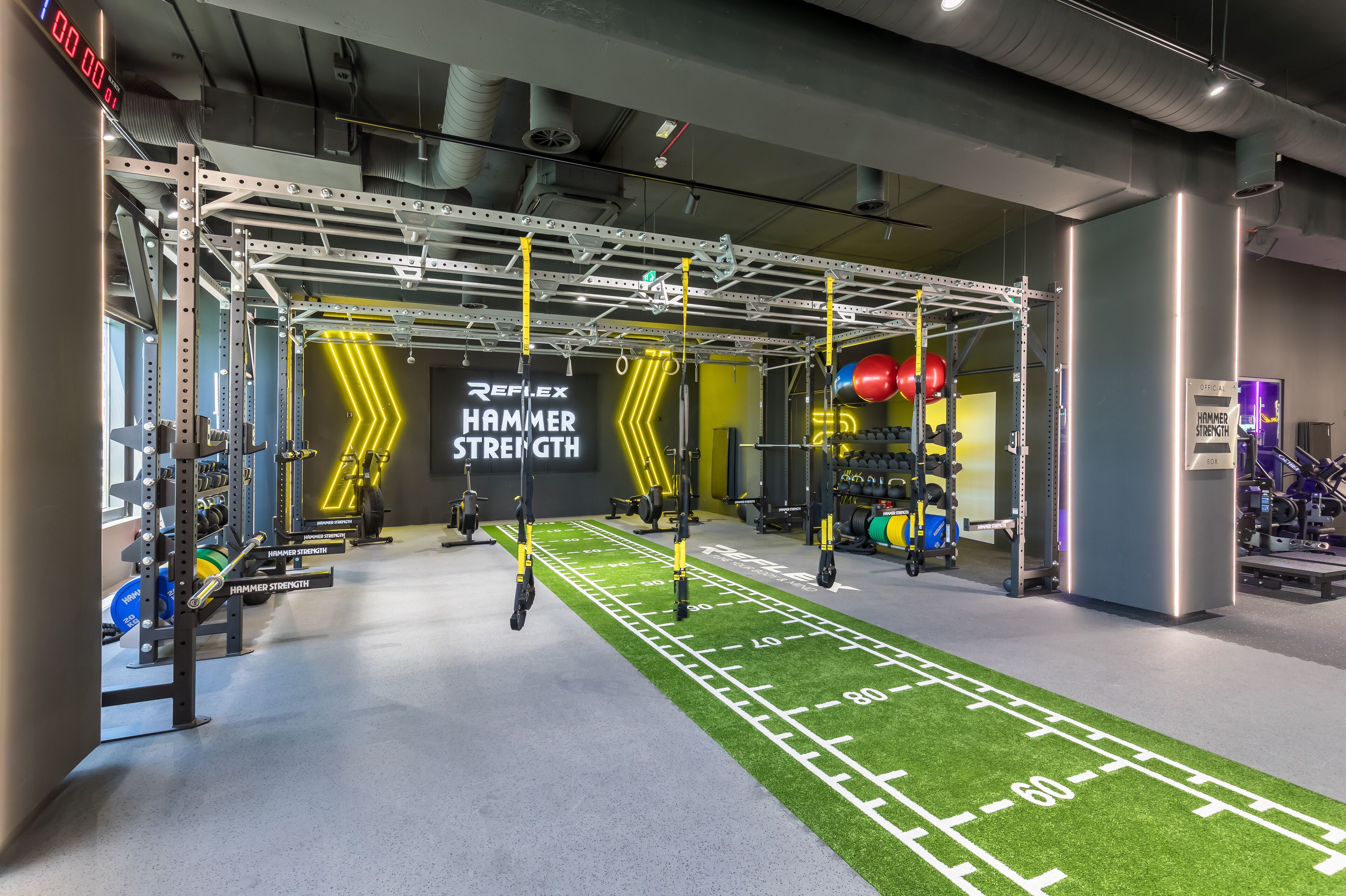 A gym with a green turf running track, weightlifting equipment, and a neon sign reading "REFLEX HAMMER STRENGTH".