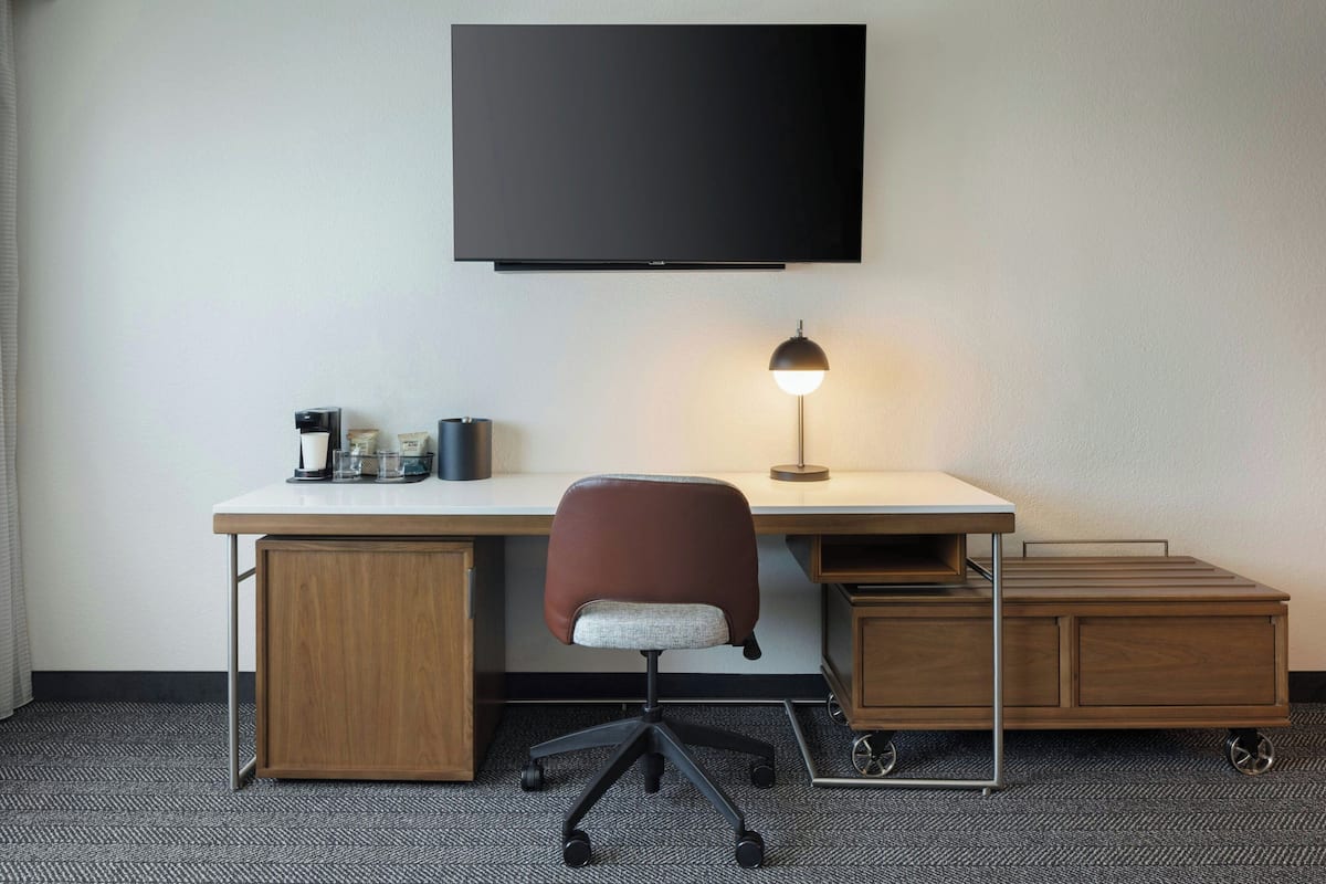 A modern hotel room with a flat-screen TV mounted on the wall, a desk with a chair, a lamp, and a small cabinet.