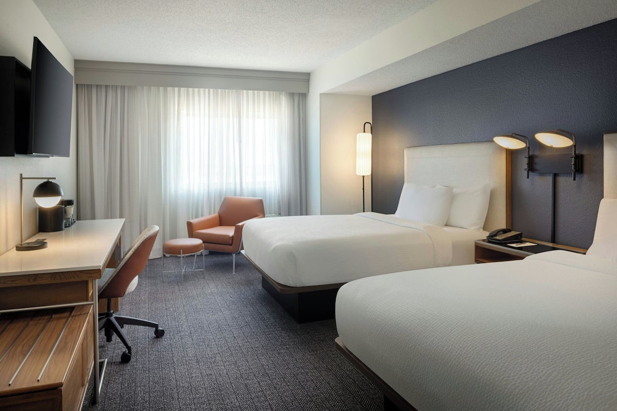 Ein Hotelzimmer mit zwei Betten, einem Schreibtisch, einem Stuhl, einem Fernseher und einem großen Fenster mit Vorhängen.