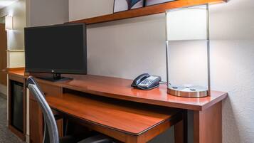 A hotel room with a wooden desk, a flat-screen TV, a telephone, and a desk lamp.