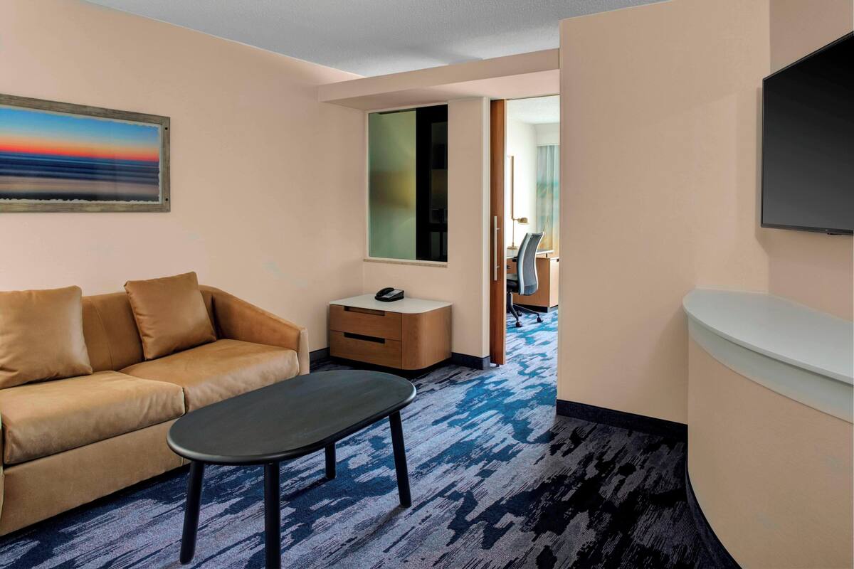 A hotel room with a sofa, a small table, a nightstand, a TV, and a large window with a view of the sunset.