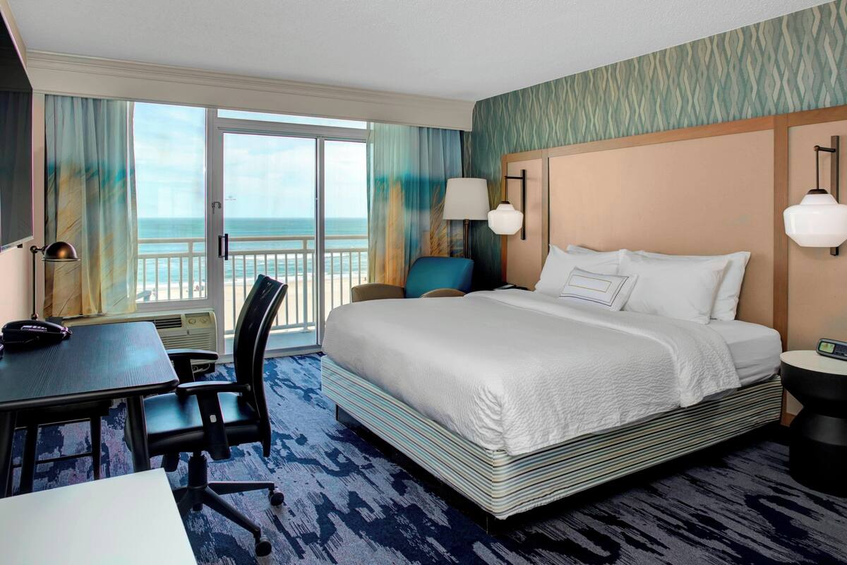 A hotel room with a large bed, a desk, a chair, a nightstand, and a view of the ocean.