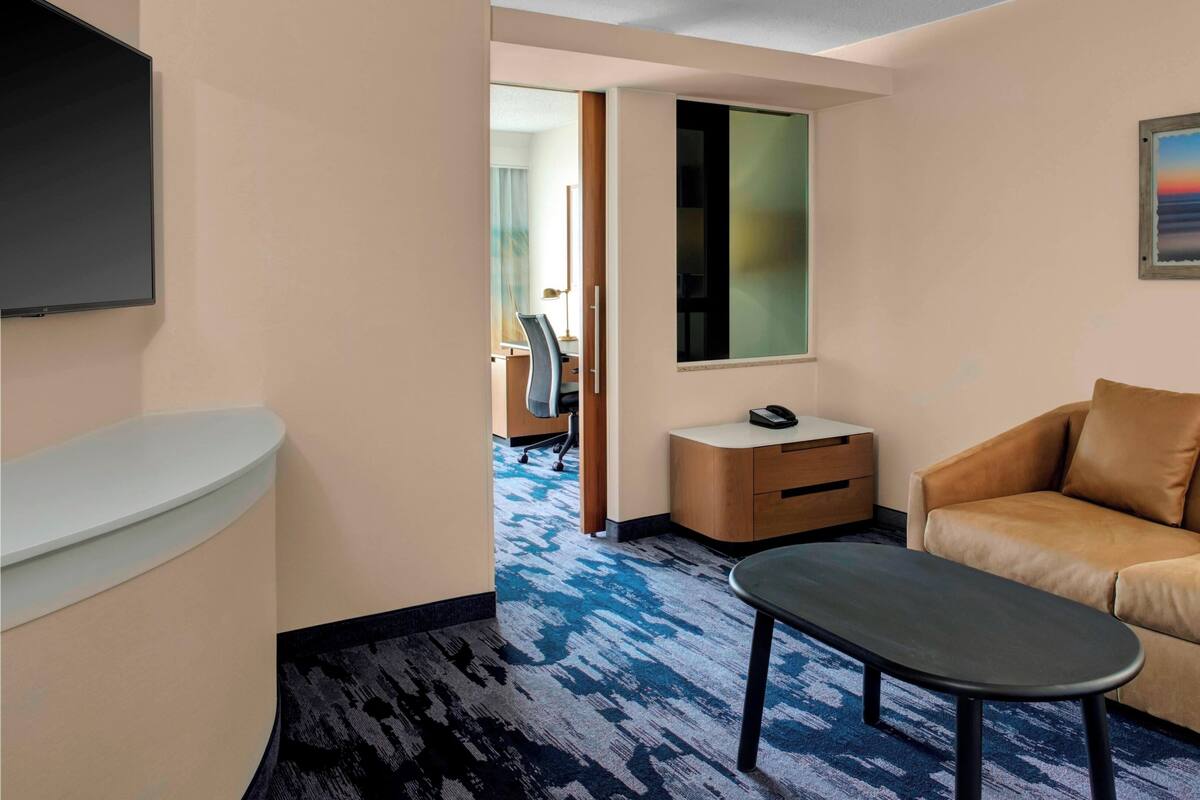 A hotel room with a sofa, a small table, a desk with a chair, a TV mounted on the wall, and a painting of a sunset.