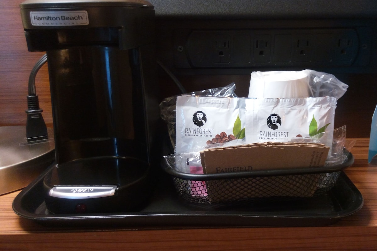 Eine Hamilton Beach Kaffeemaschine auf einer schwarzen Ablage, mit einer Kaffeekapsel und einer Packung Rainforest Premium Select Kaffee.
