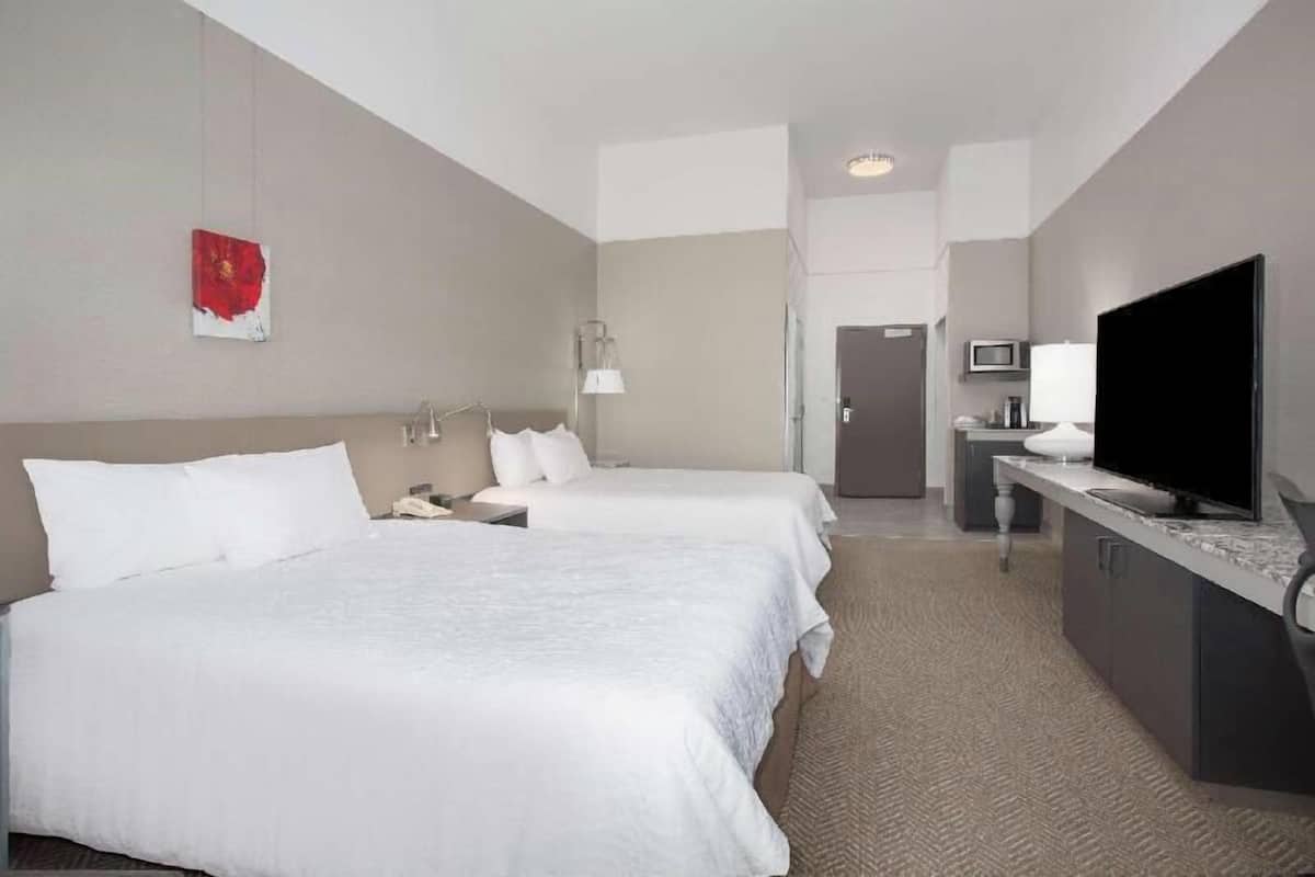 A hotel room with two beds, a desk with a TV, a lamp, and a small kitchenette.