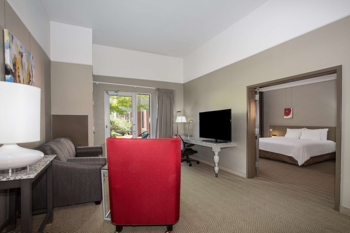 A hotel room with a bed, a desk with a TV, a red chair, a sofa, and a window with curtains.