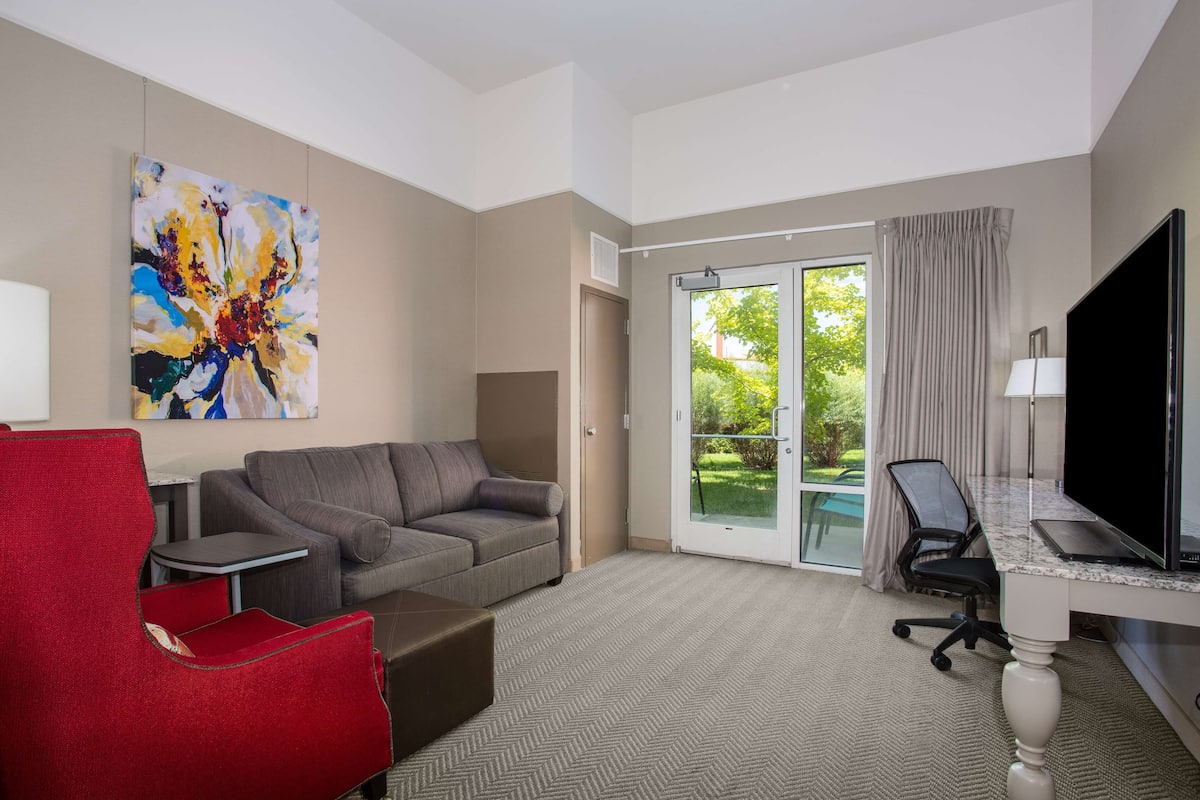 A hotel room with a gray sofa, a red armchair, a desk with a computer, a TV, and a large window with a view of greenery.