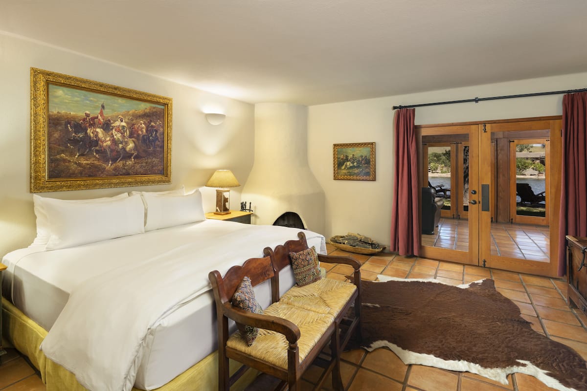 Habitación de hotel con una cama grande, un banco de madera, un tapiz de cuero de vaca, un cuadro y vista a una piscina por la puerta.