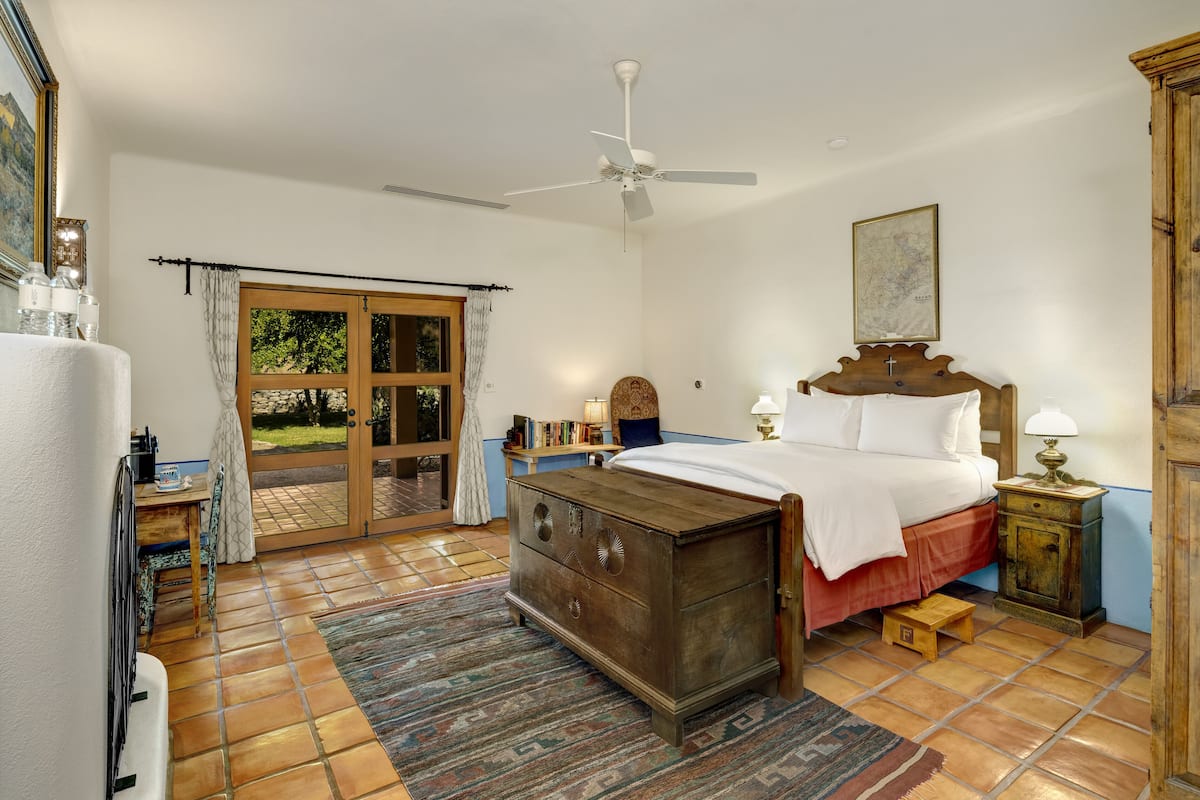 Un dormitorio con una cama, un aparador de madera, una mesita de noche, un ventilador de techo y un ventanal con vistas al verdor.