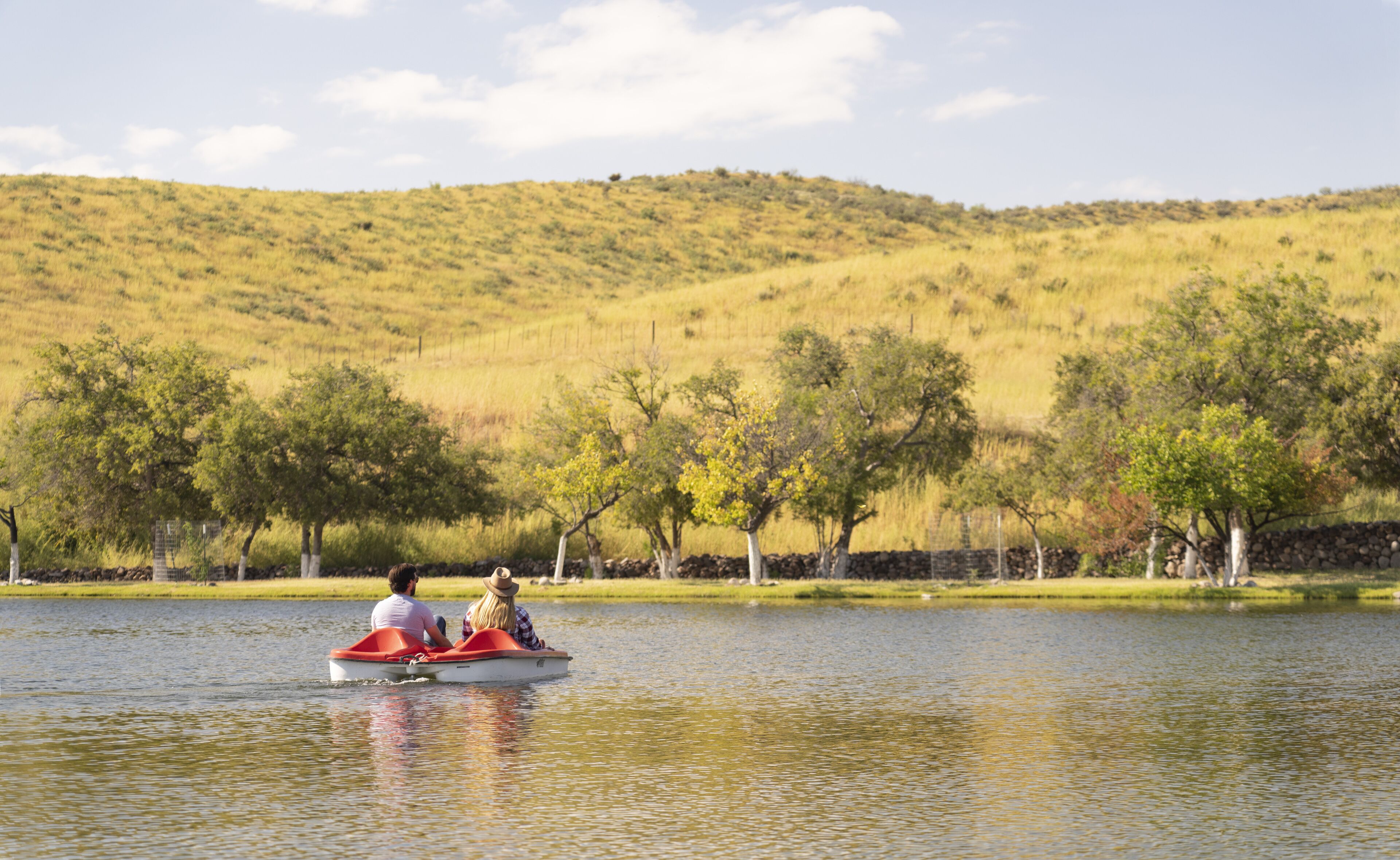 A couple in a small boat on a calm lake with a backdrop of trees and hills.