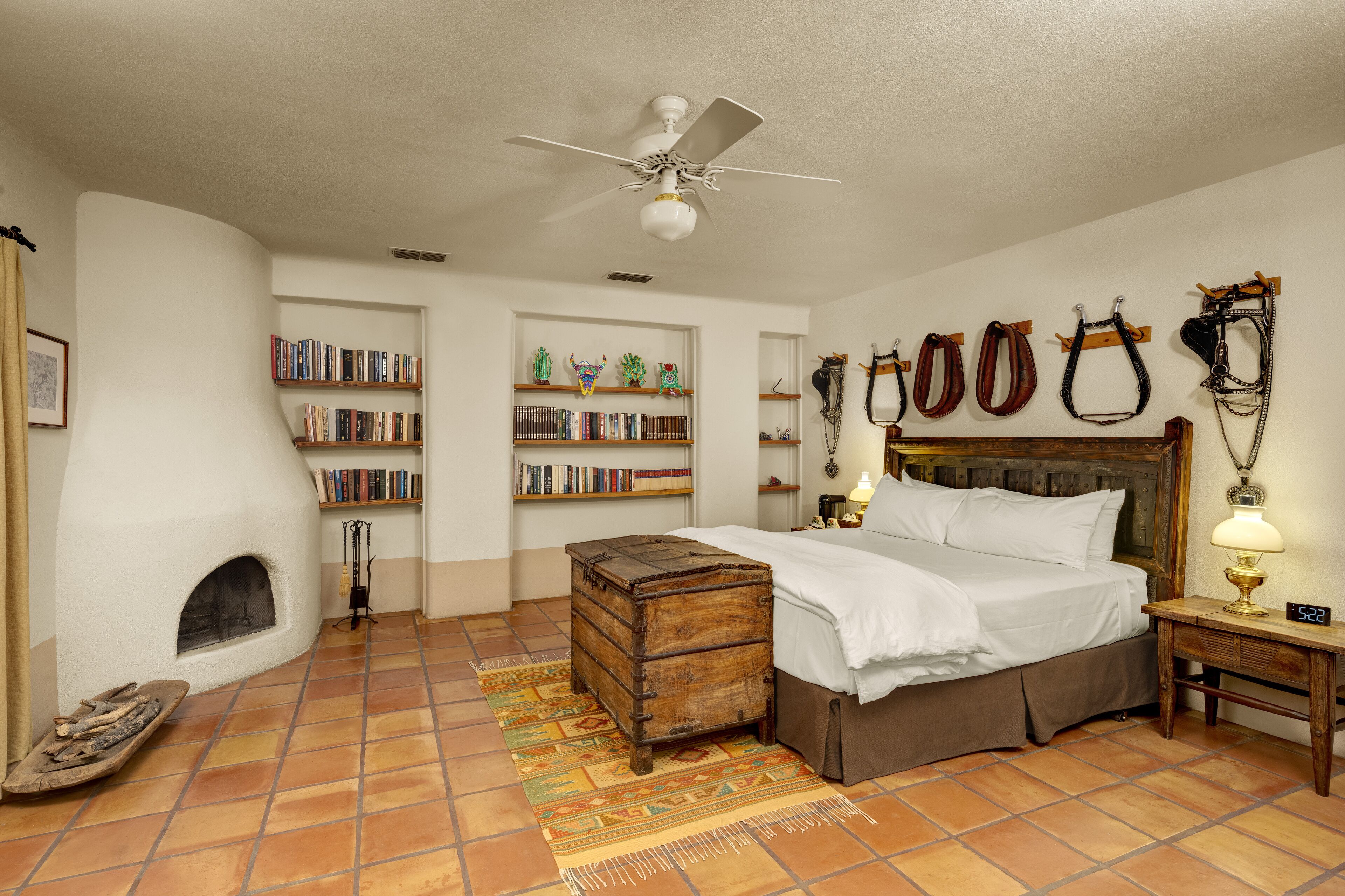A bedroom with a bed, a wooden chest, a fireplace, bookshelves, and horse tack on the wall.