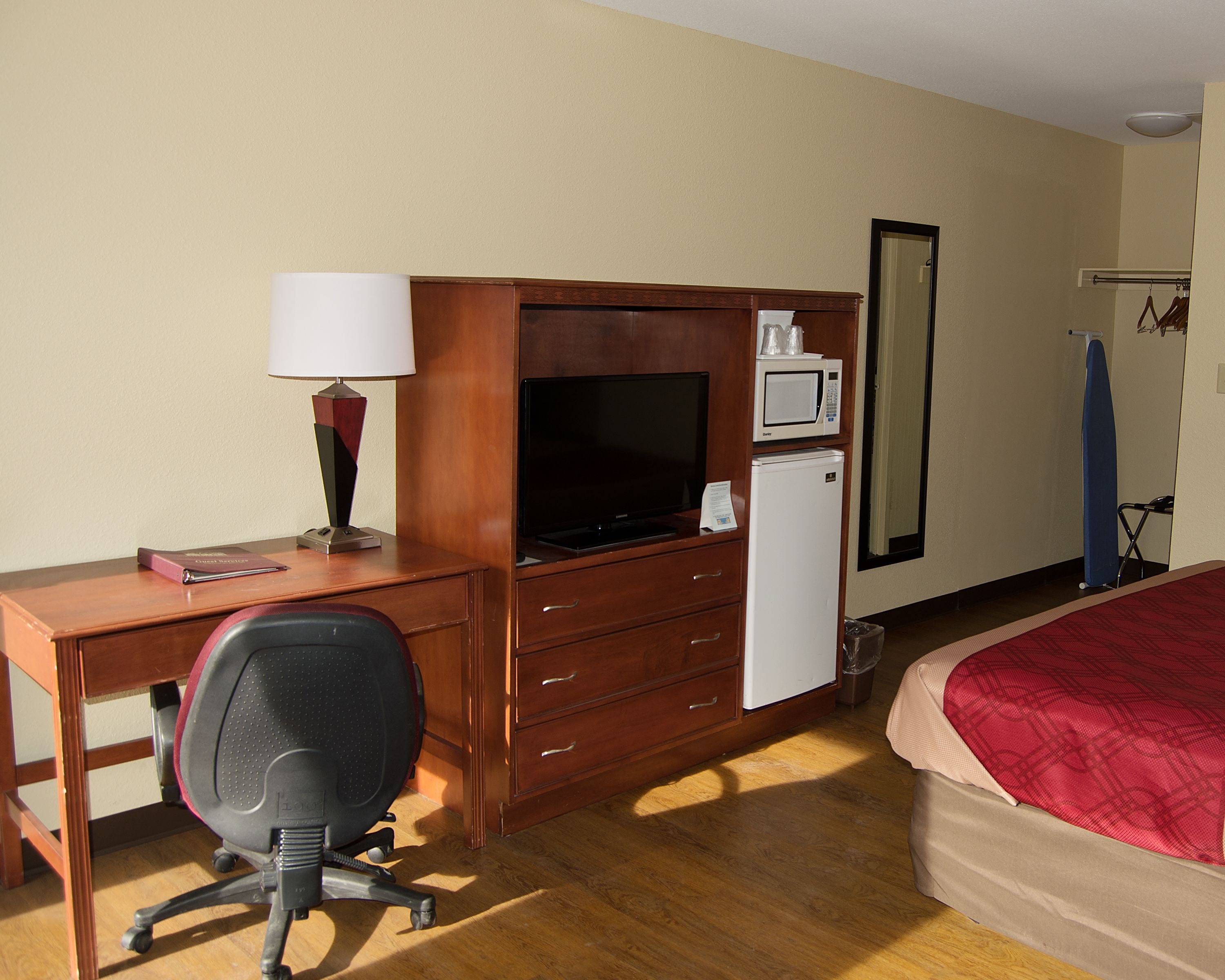 A hotel room with a wooden desk, a television, a microwave, a refrigerator, a chair, a bed with a red blanket, and a mirror.