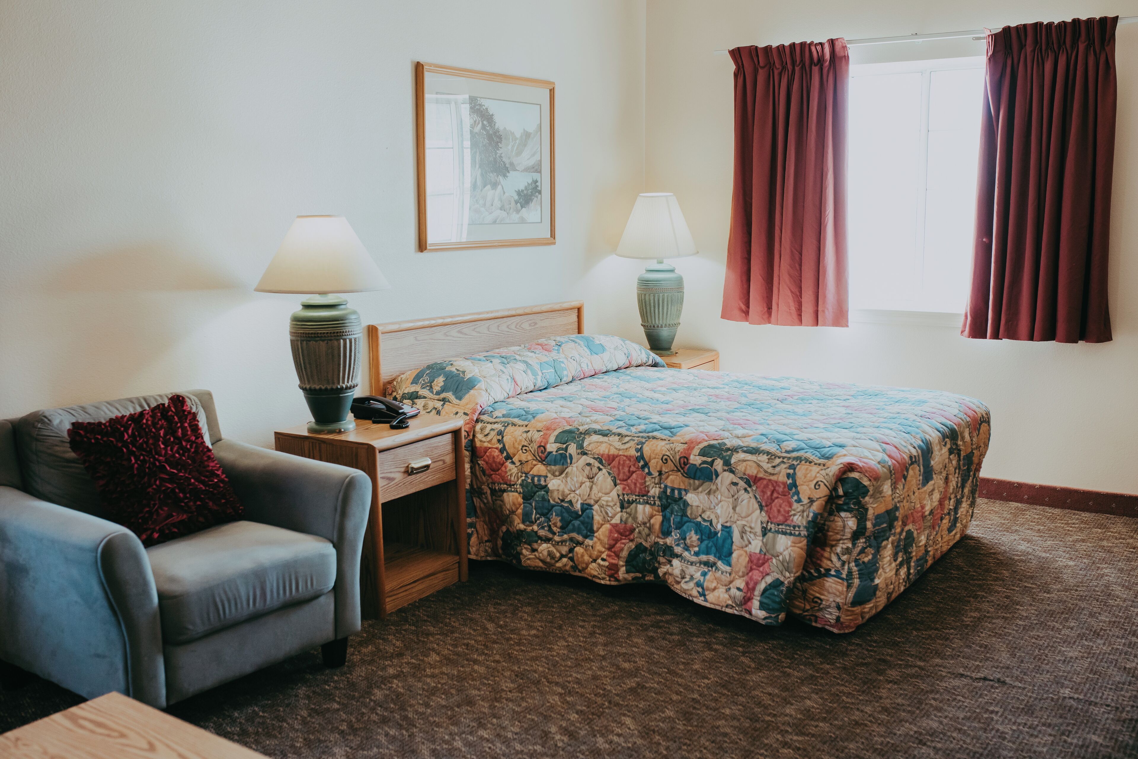 A bedroom with a bed, a chair, two bedside tables with lamps, a framed picture on the wall, and a window with curtains.