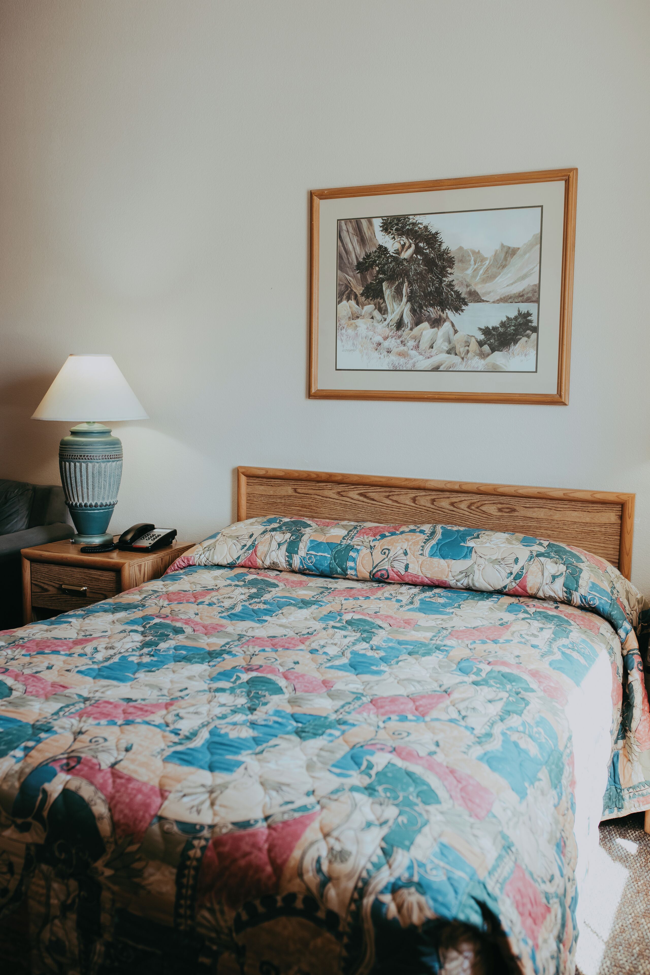 Una cama bien hecha con una colcha floral, un cabecero de madera, una lámpara de noche, un cuadro en la pared y una mesita de noche con un teléfono y una lámpara.