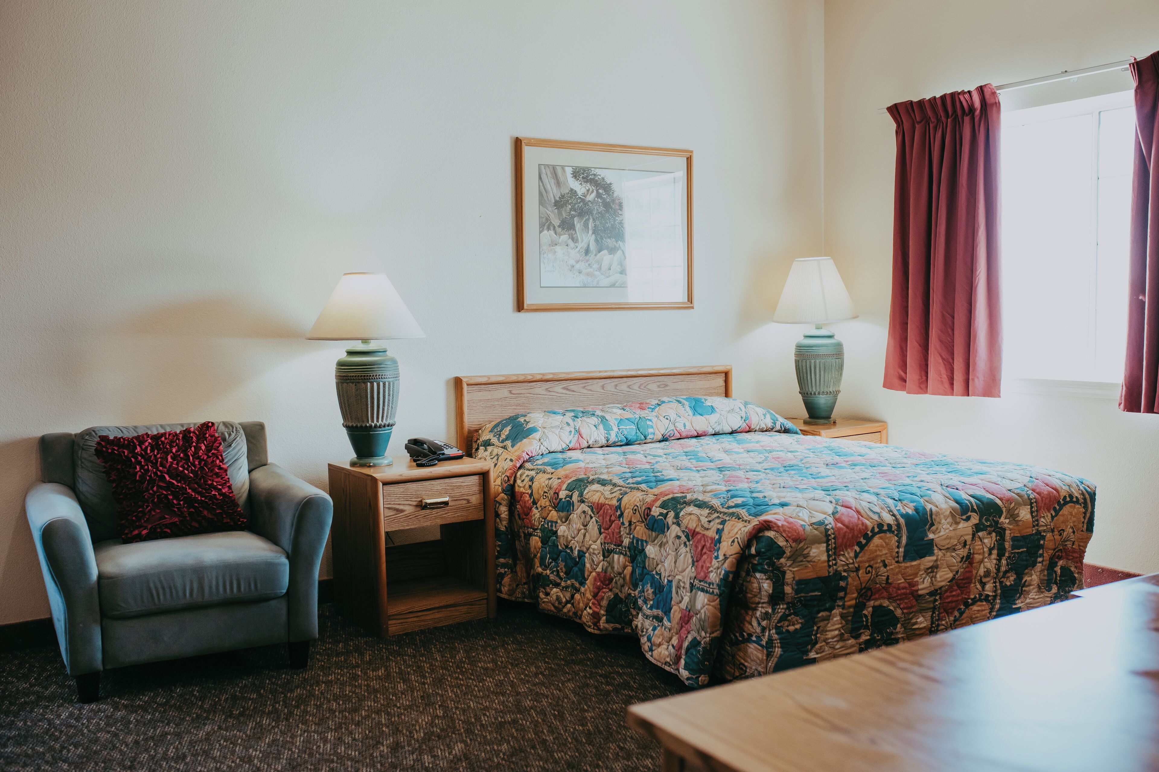 A hotel room with a bed, a chair, a nightstand, a lamp, and a framed picture on the wall.