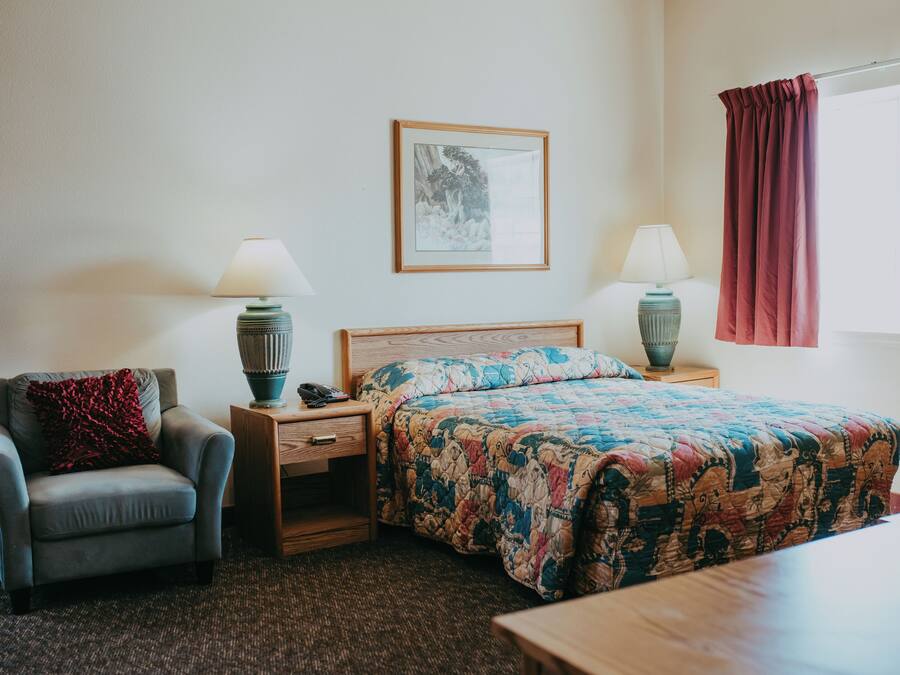 Una habitación de hotel con cama, una silla, una mesita de noche, una lámpara y un cuadro en la pared.