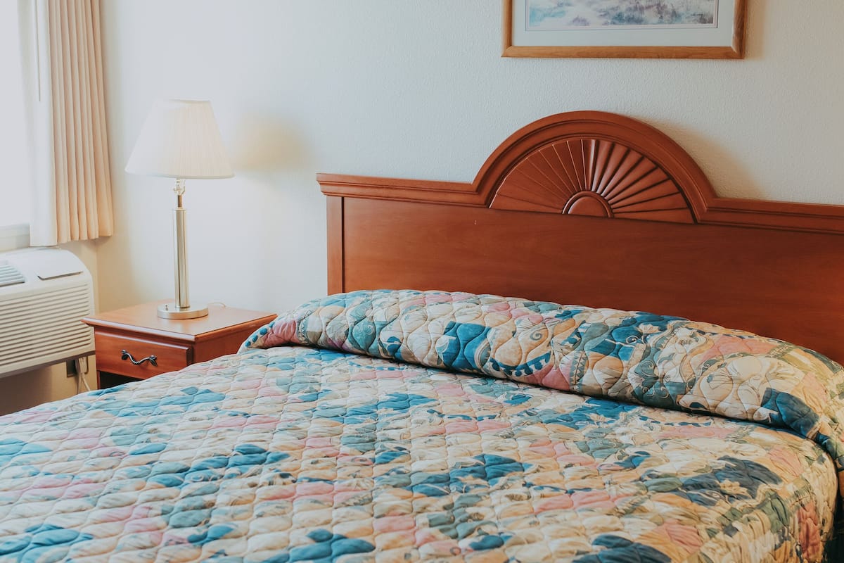 Een hotelkamer met een houten hoofdbord, een geborduurde spreidekbed, een bedlampje, een ingelijst kunstwerk aan de muur en een raam met gordijnen.