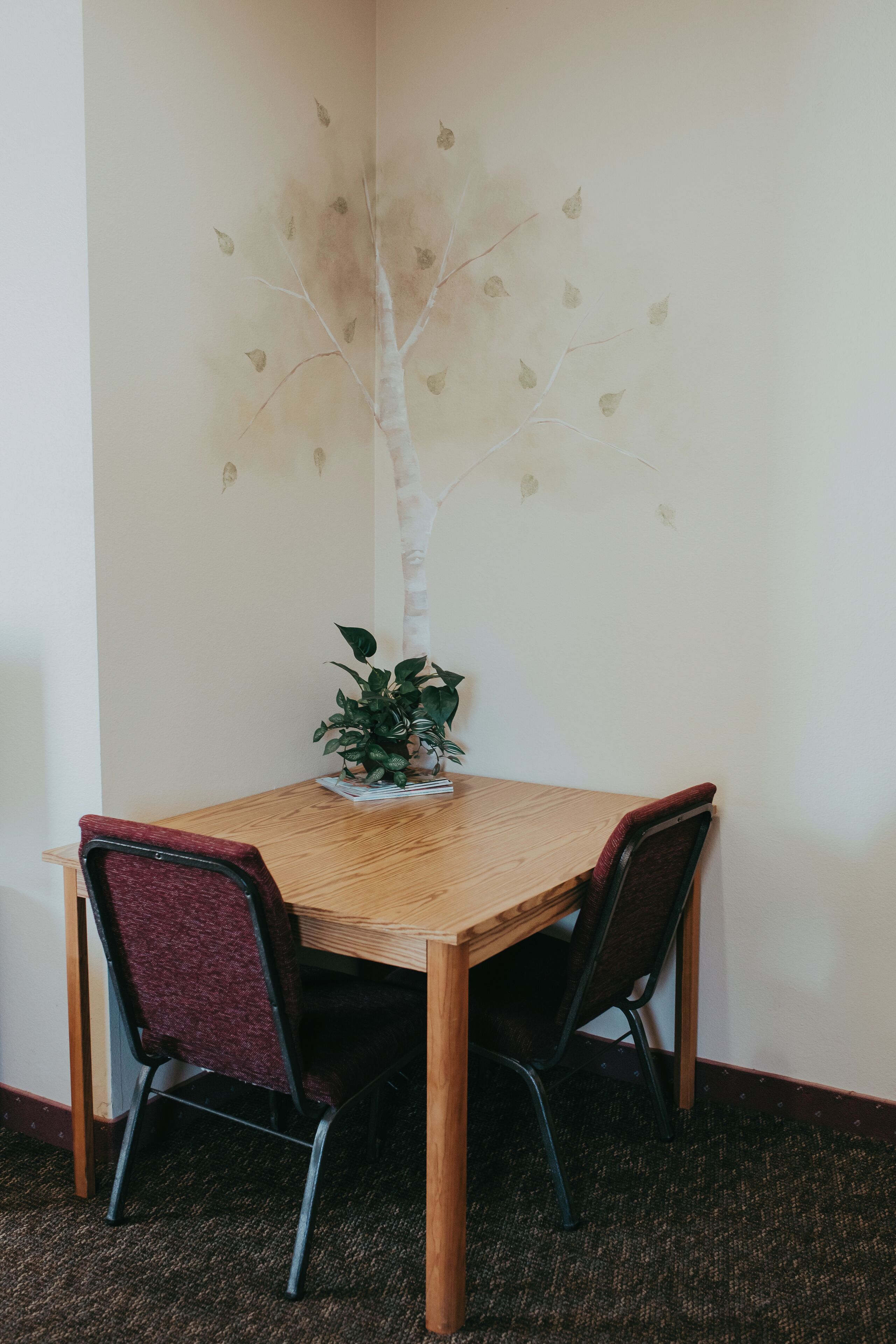 A dining area with a wooden table, two chairs, a potted plant, and a wall mural of a tree.