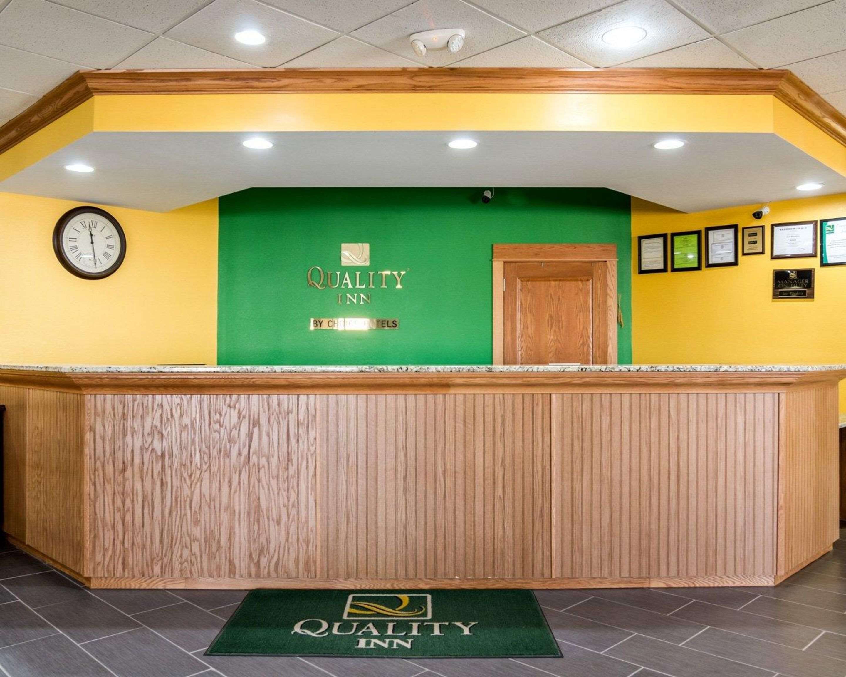 Zona de recepción con mostrador de madera, un reloj y una pared verde con el logotipo de Quality Inn.