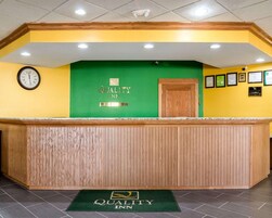 Zona de recepción con mostrador de madera, un reloj y una pared verde con el logotipo de Quality Inn.