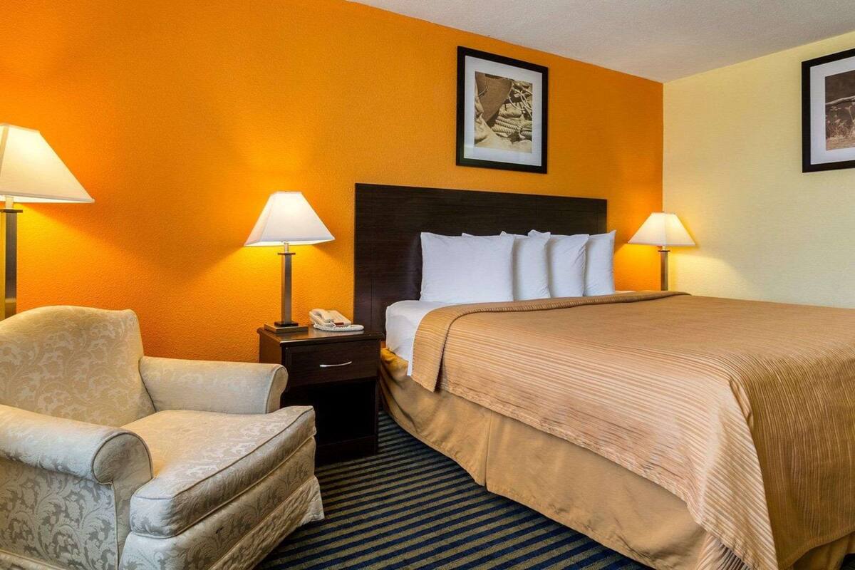 Quarto de hotel com cama, uma cadeira, duas mesas de cabeceira com luminárias e um quadro na parede.