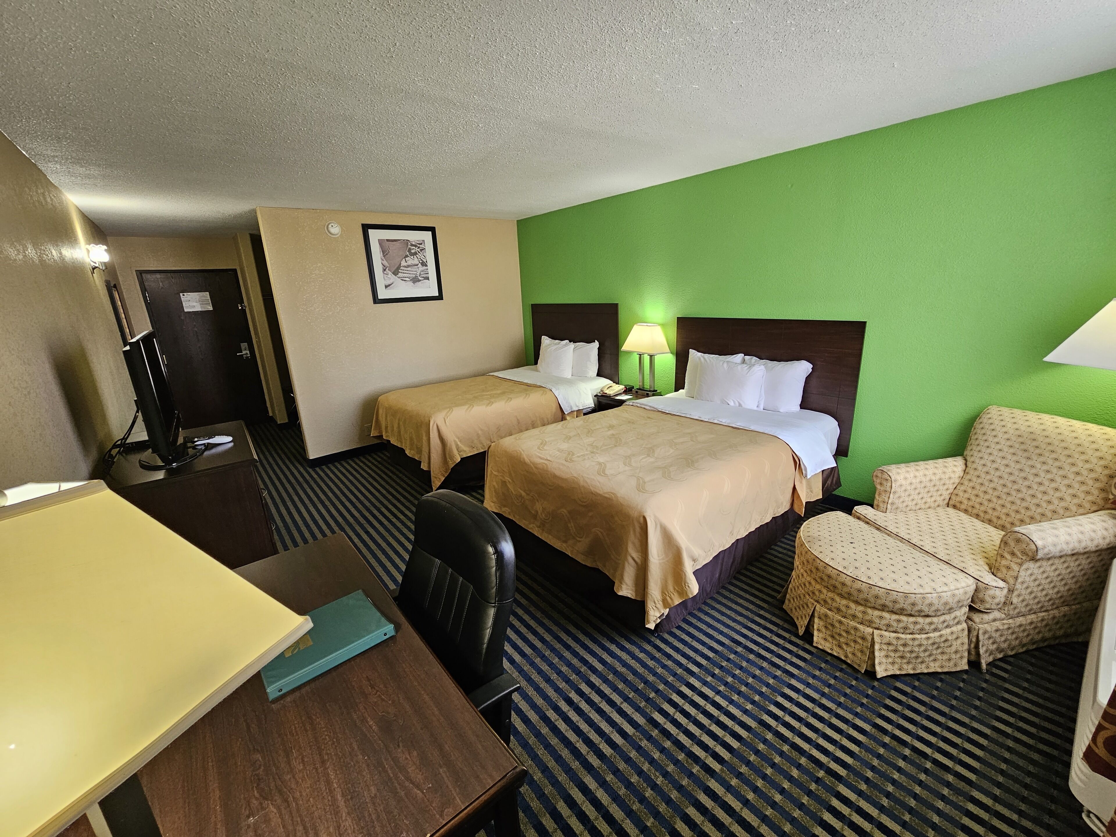 A hotel room with two beds, a desk, a chair, and a lamp.