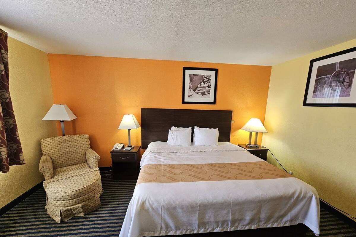 Quarto de hotel com cama, duas luminárias de cabeceira, uma cadeira, mesa de cabeceira e um quadro na parede.