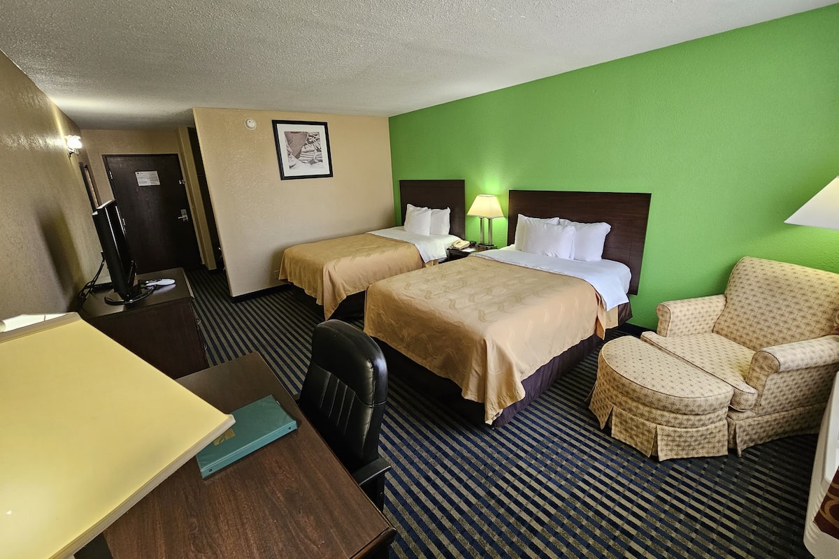 Quarto de hotel com duas camas, uma escrivaninha, uma cadeira, um abajur e um quadro na parede.