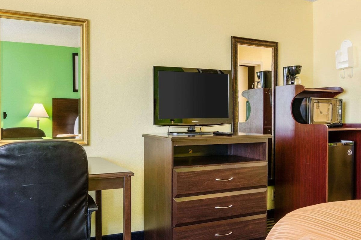 Quarto de hotel com televisão, penteadeira de madeira com gavetas, escrivaninha com abajur, cama e micro-ondas.