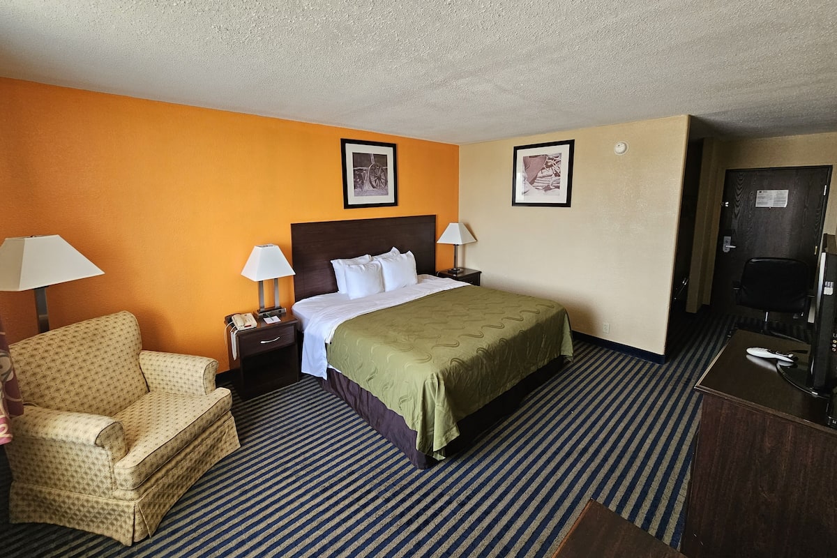 Quarto de hotel com cama, duas mesas de cabeceira, uma cadeira, escrivaninha e porta.