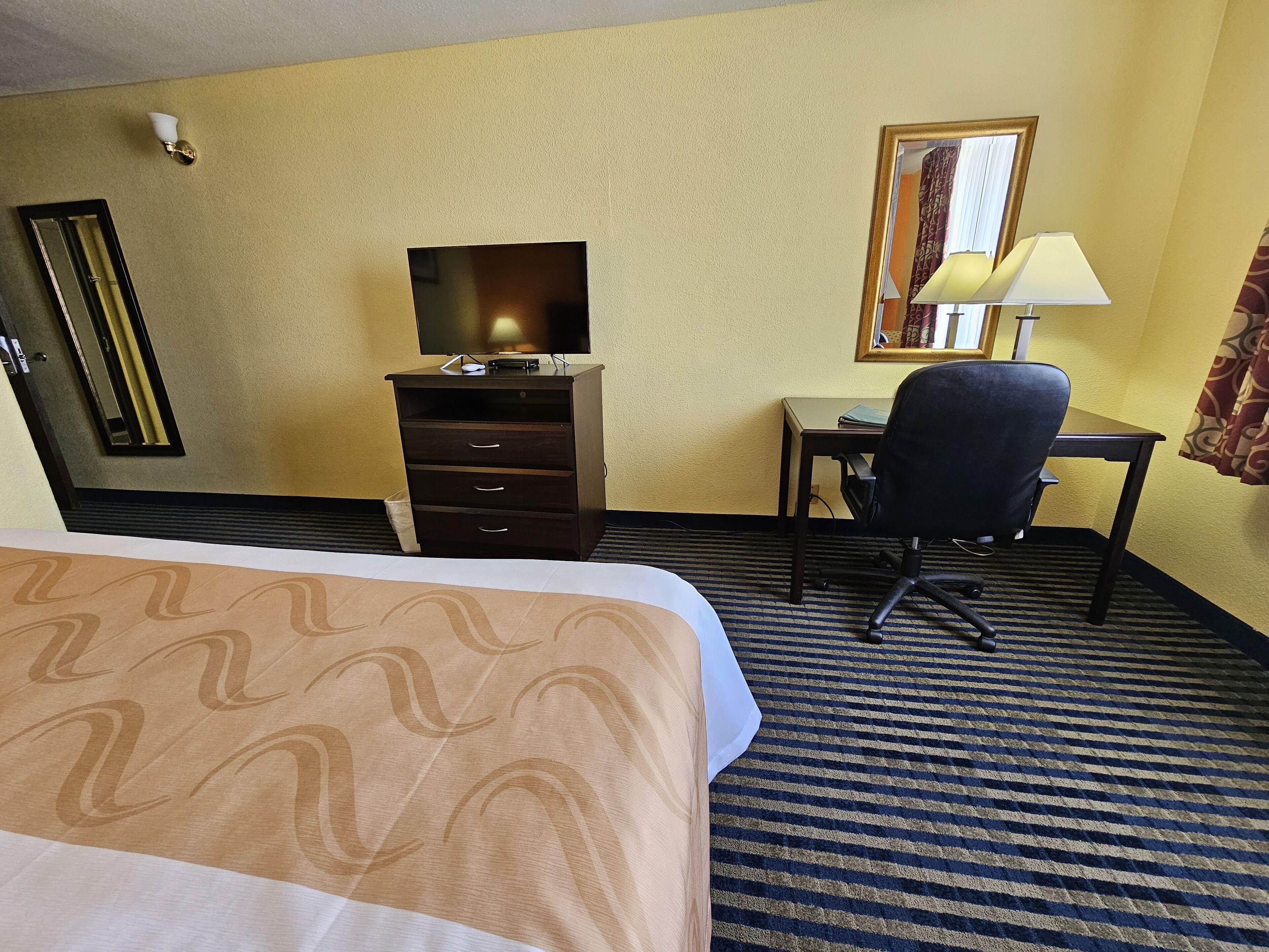 A hotel room with a bed, a desk, a chair, a television, and a lamp.