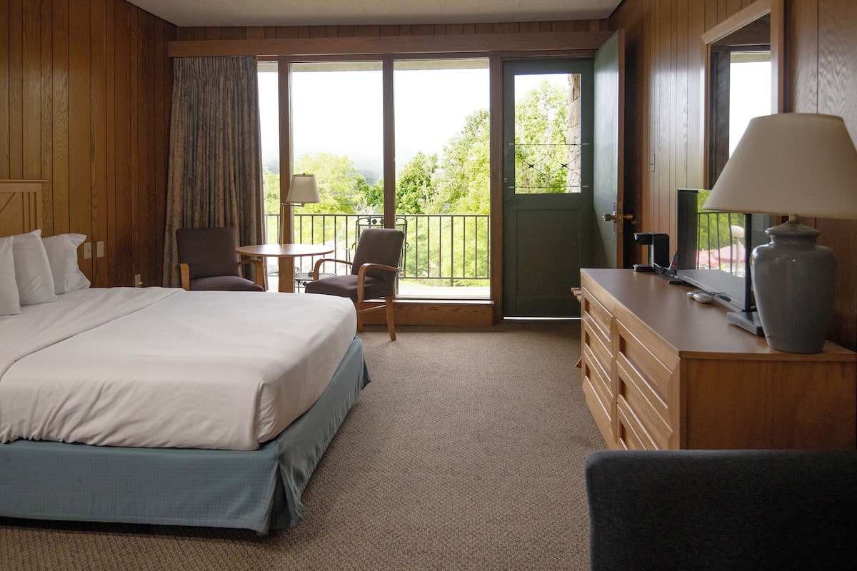 A hotel room with a bed, a desk with a lamp, a chair, and a view of the outdoors through a door.