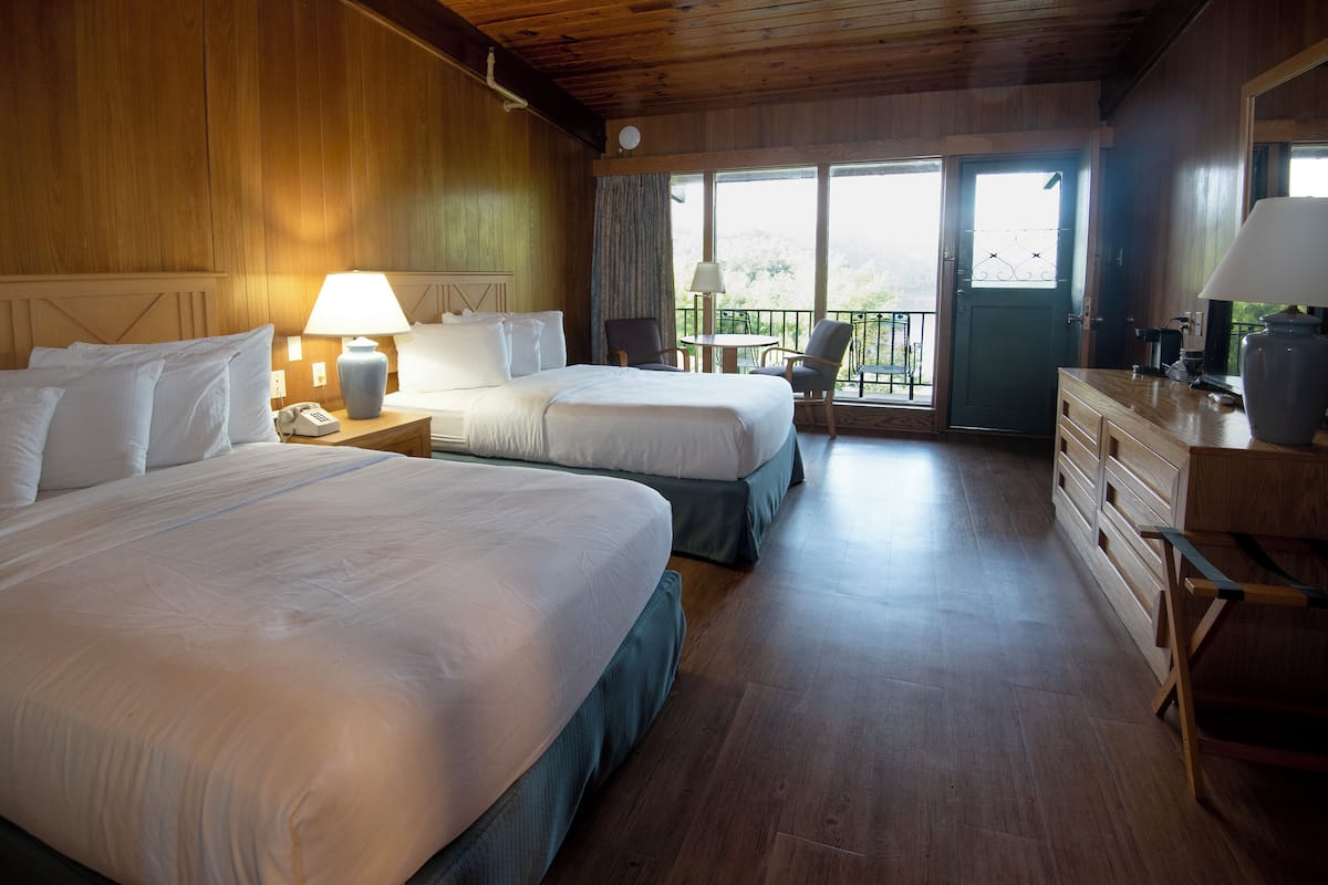 A hotel room with two beds, a wooden floor, a balcony with a view, and a dresser with a lamp.