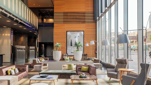 A modern lobby with a variety of seating options, including sofas, chairs, and ottomans. There is a large abstract painting on the wall, and the space is well-lit with natural light from large windows. - Leonardo Royal Hotel Barcelona Forum (Barcelona)