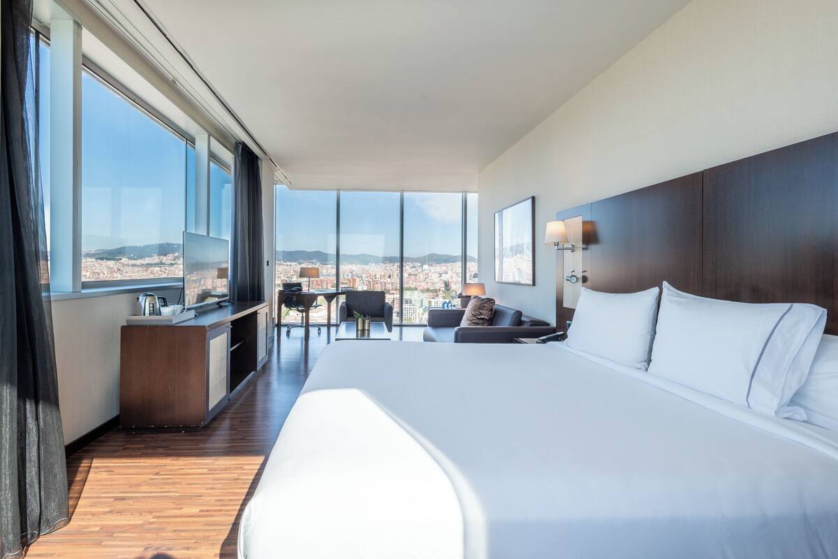 A modern hotel room with a large bed, a desk, a chair, and a view of the city.