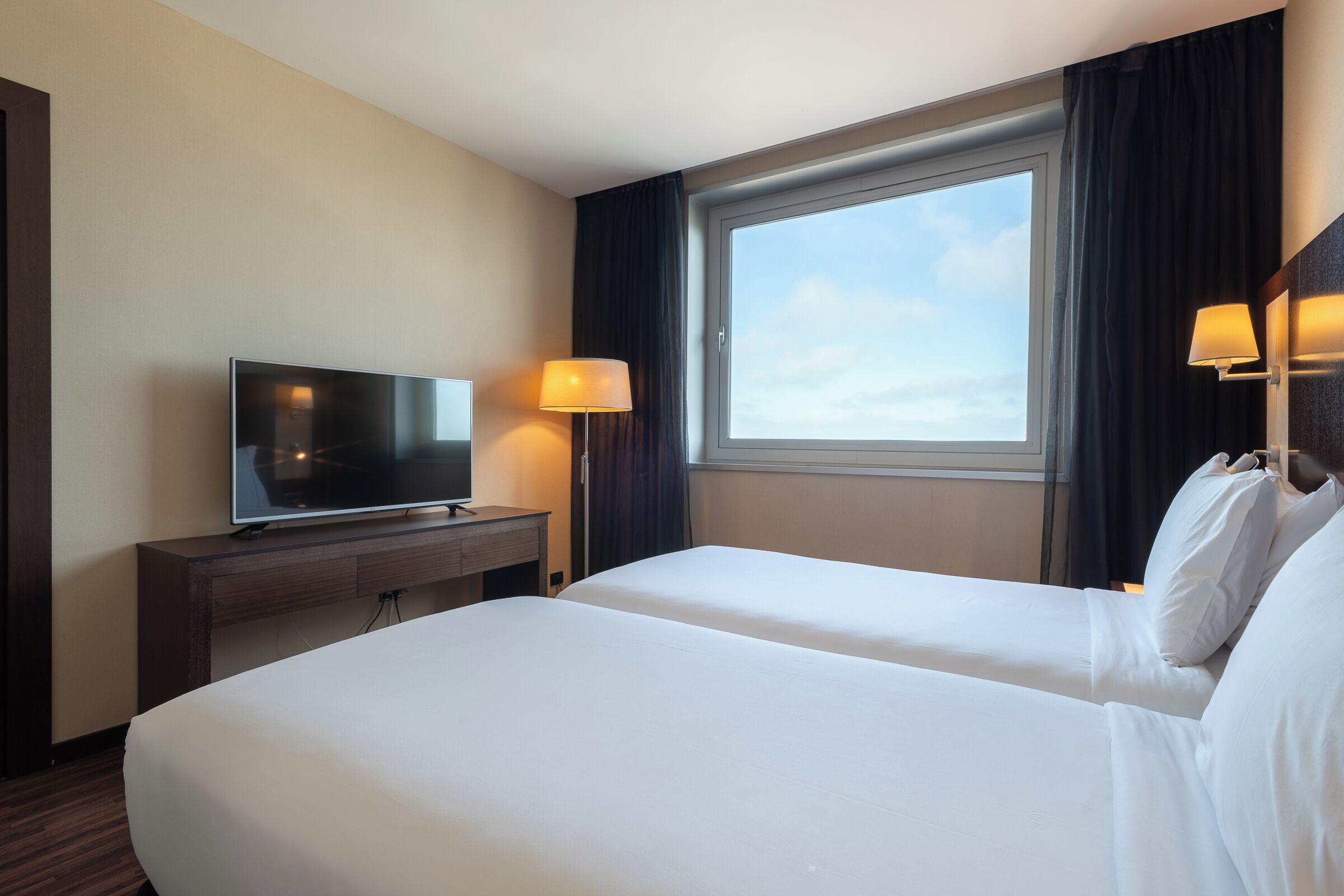 A hotel room with two beds, a TV on a wooden cabinet, and a window with curtains.