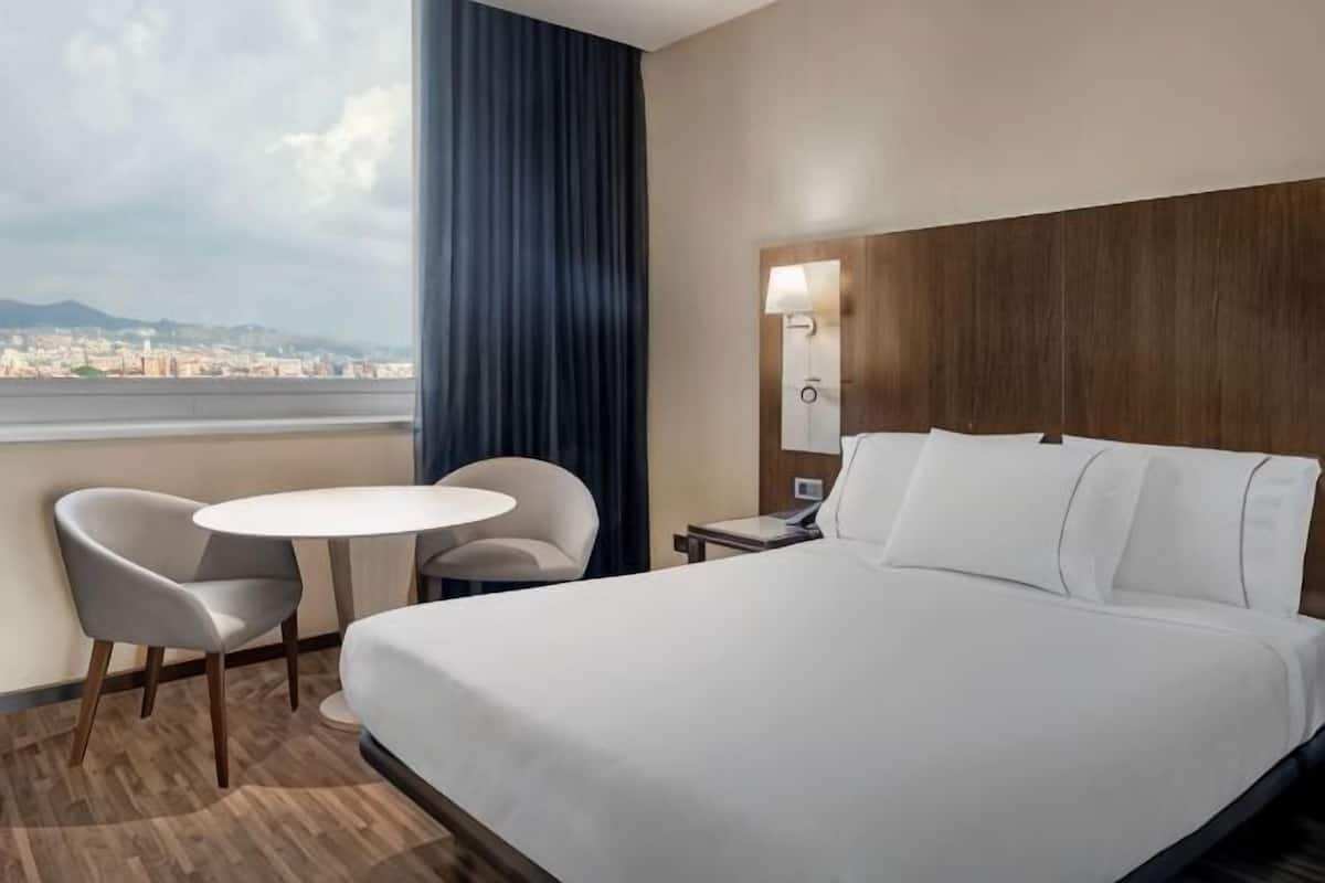 A hotel room with a bed, a round table, two chairs, a nightstand, and a view of a cityscape.