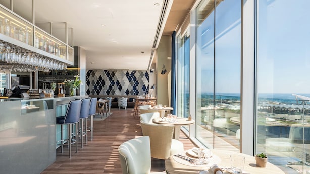 A modern bar area with a high bar counter, seating with bar stools, and a view of the cityscape through large windows. - Leonardo Royal Hotel Barcelona Forum (Barcelona)