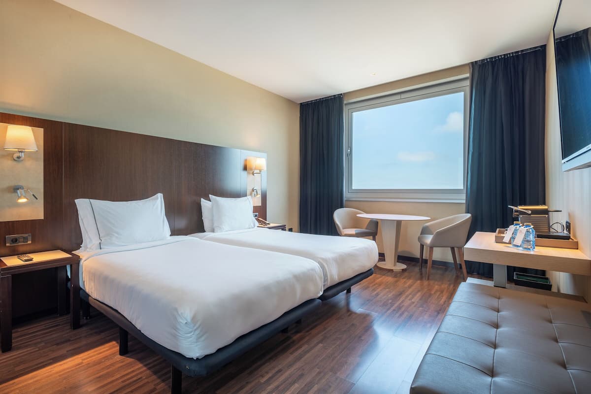 A hotel room with two beds, a small round table, a chair, a sofa, and a window with curtains.