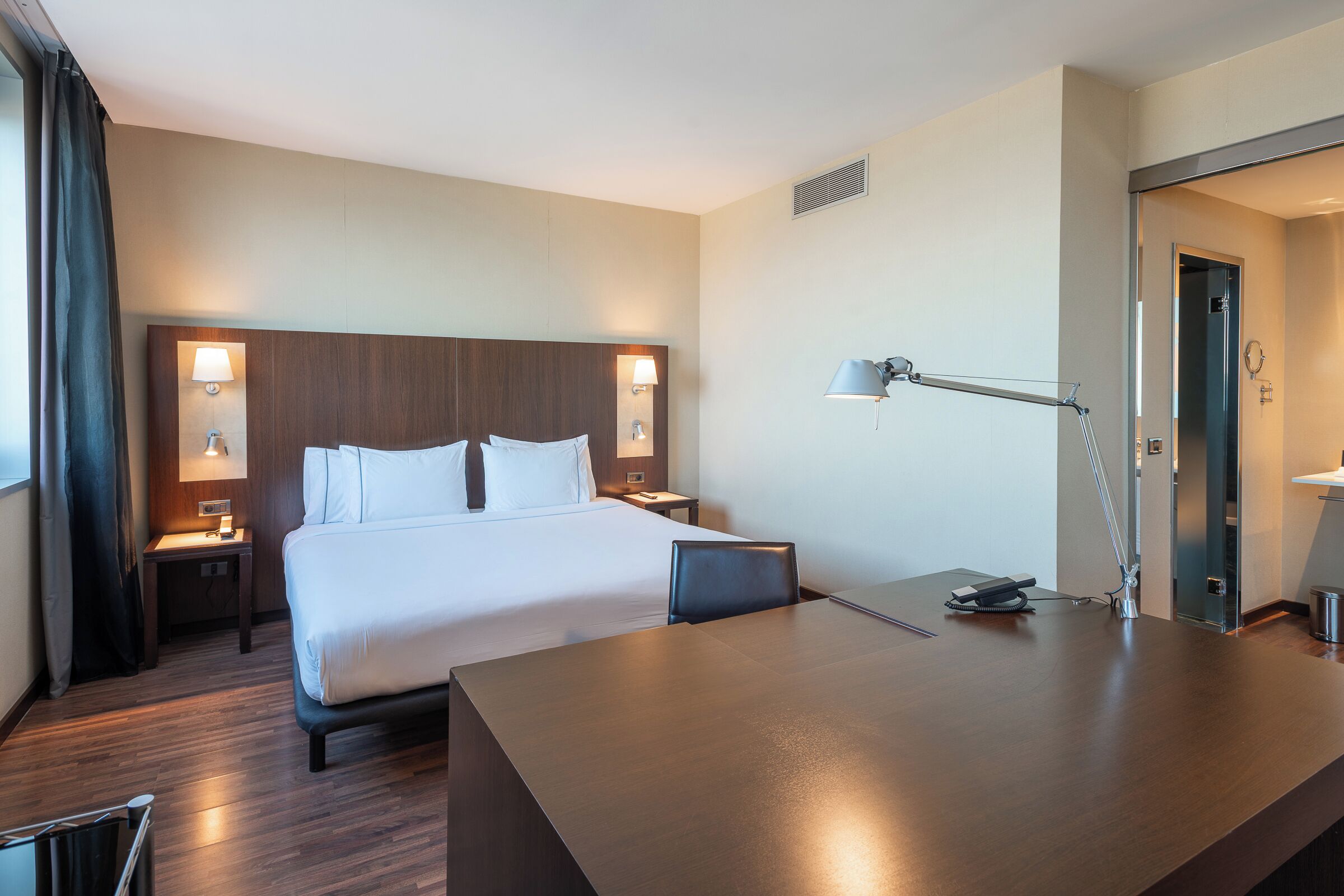 A hotel room with a large bed, a desk, and a chair. There are two bedside tables with lamps and a mirror.