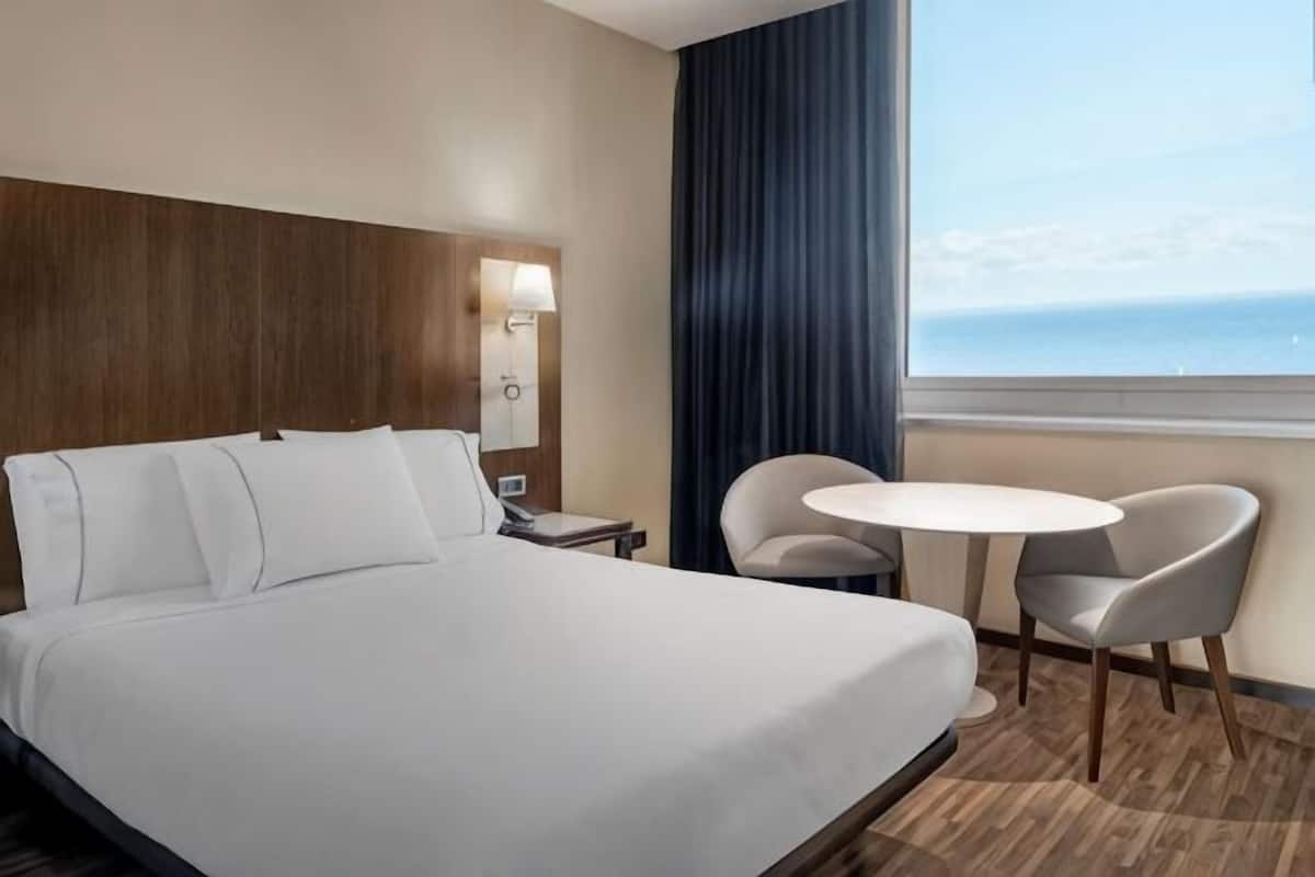 A hotel room with a large bed, a bedside table, a chair, a small round table, and a view of the ocean.