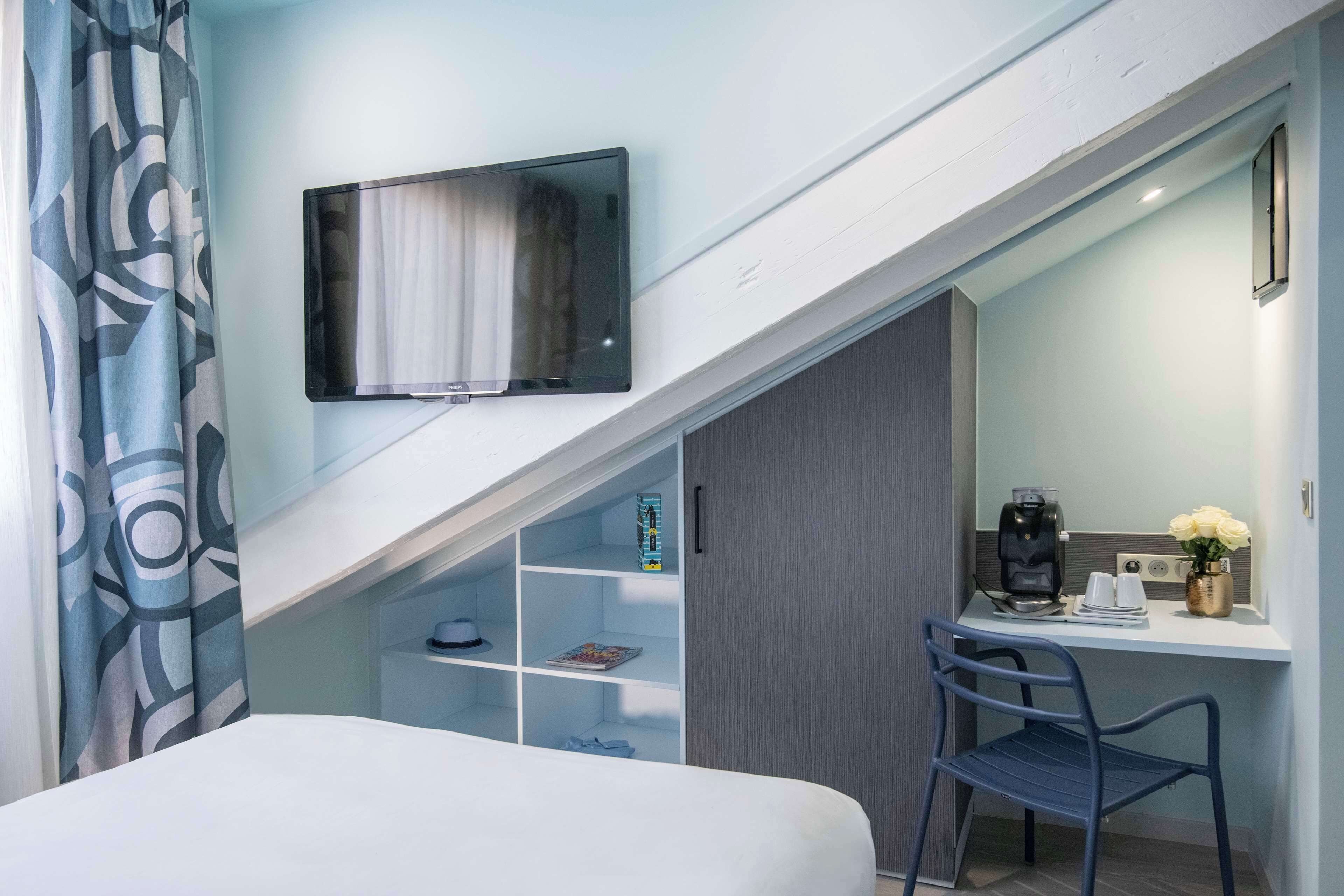 Una habitación de hotel con cama, escritorio con silla, televisor, cafetera y estante con libros y un sombrero.