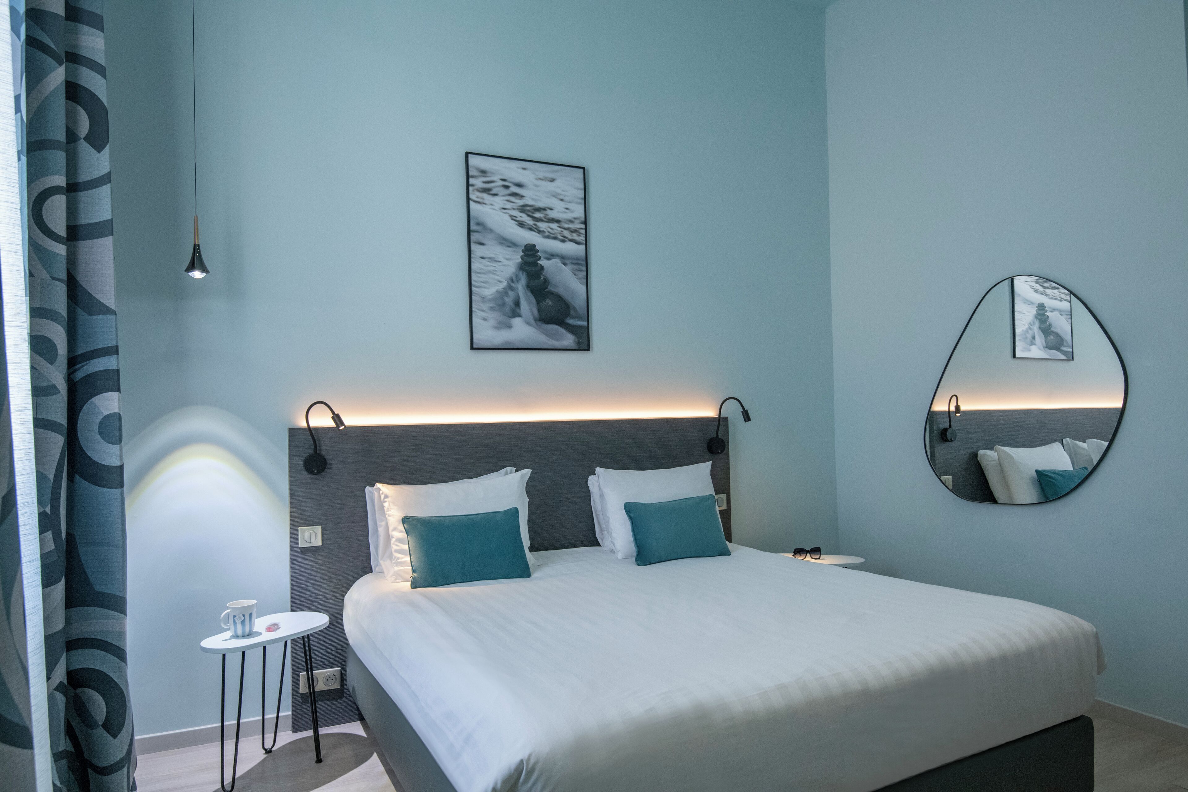 Una habitación de hotel moderna con una cama grande, una mesita de noche pequeña, un espejo fijado a la pared y una imagen de un paisaje nevado.