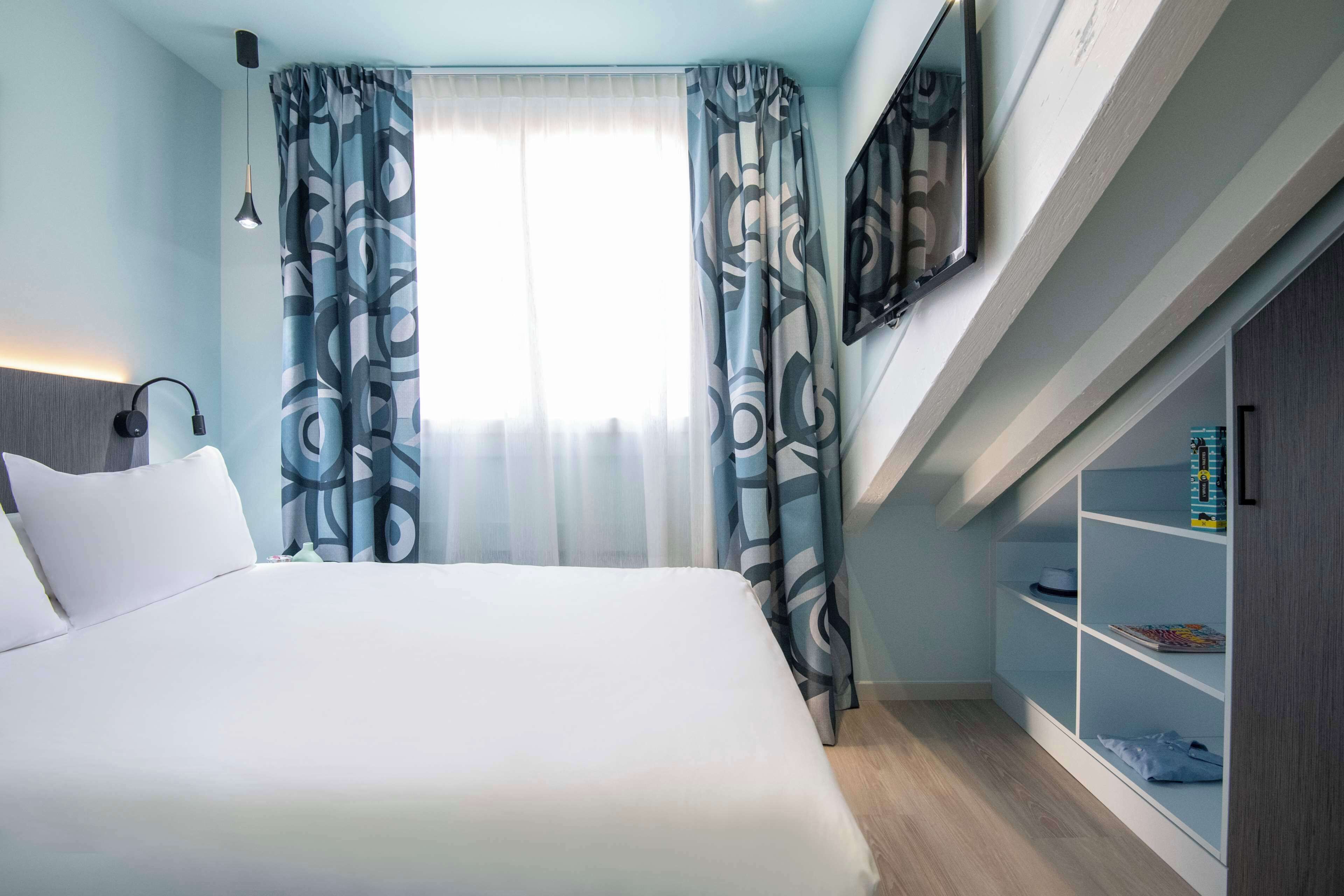 A modern hotel room with a large bed, a desk, a bookshelf, and a window with patterned curtains.